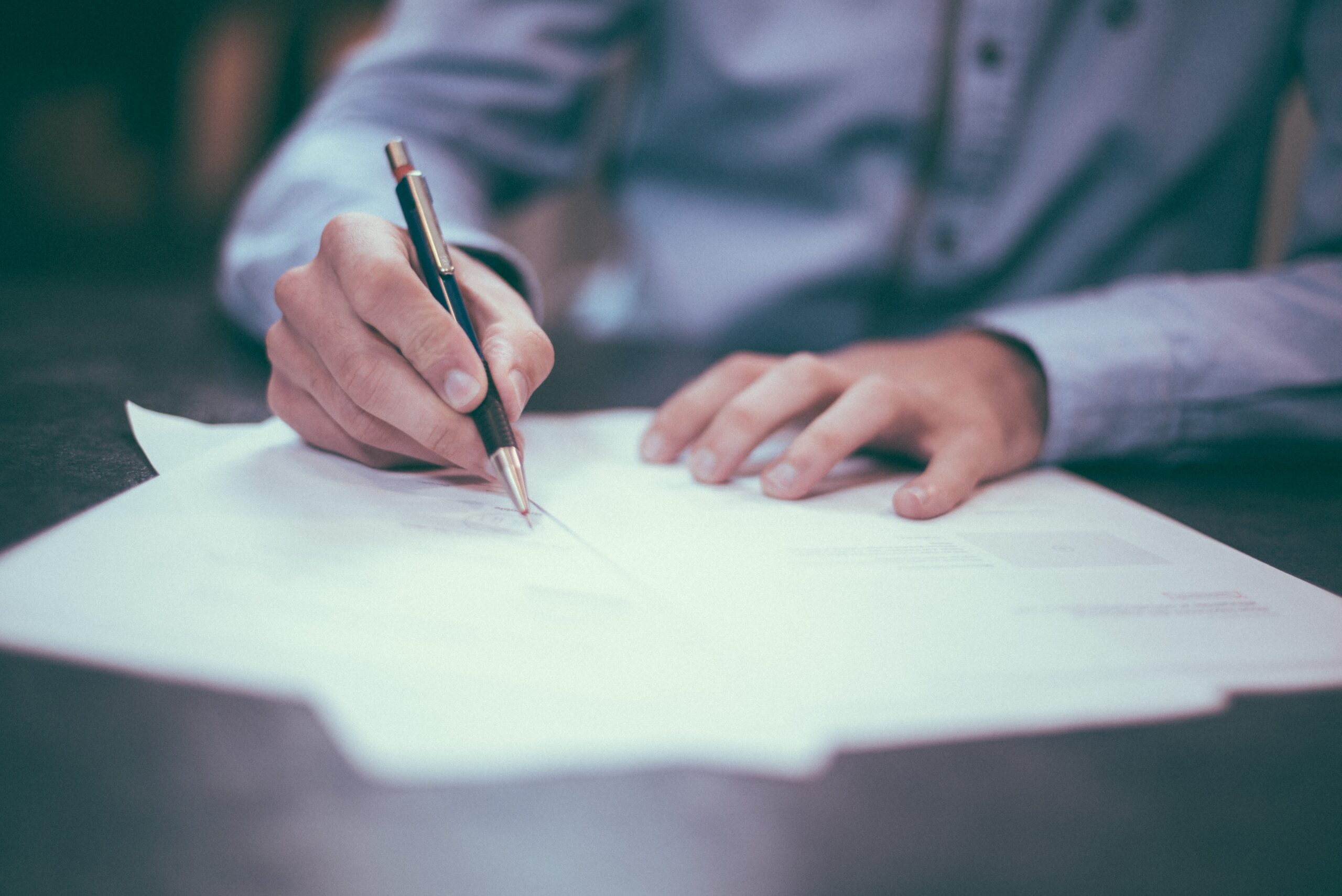 Image of a person signing 3 formal documents with a pen.